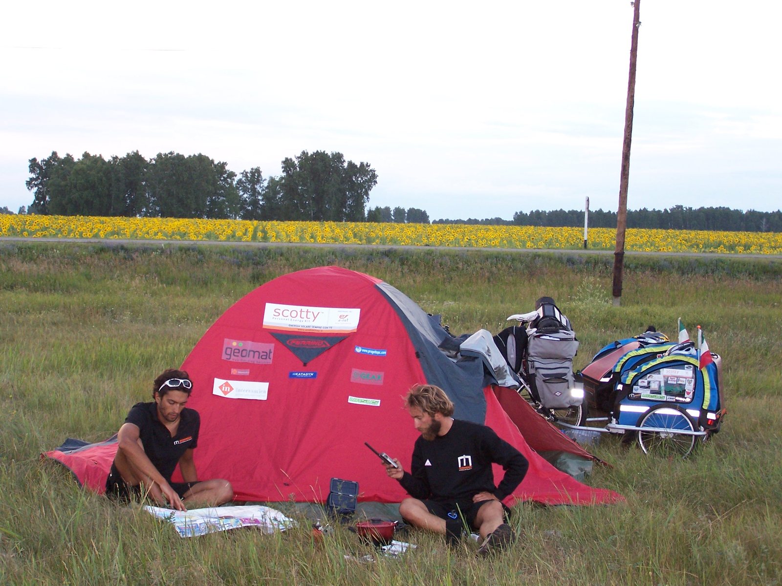 Il campo tenda tappezzato dei loghi sponsor — Scotty energia solare, Geomat, Intermatica — con Lenny al telefono satellitare
